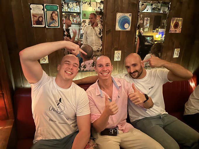 Three smiling people sitting together on a red bench; two men in white shirts give thumbs up while the woman in a pink shirt points at them.
