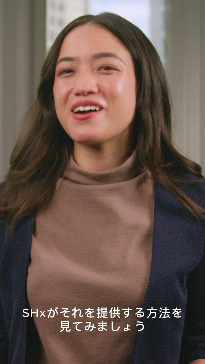 The image features a woman with long brown hair wearing a brown sweater, smiling at the camera while looking directly into it. The background of the photo shows a window, suggesting that she might be indoors or in an office setting. There are no texts or other objects visible in the image.