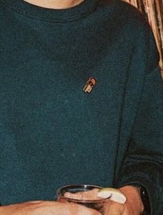 A person wearing a teal crewneck sweater with a small embroidered logo on the chest holds a wine glass, the muted tones and grainy quality evoking a cozy night in—fittingly captured as a "tinofied" moment.
