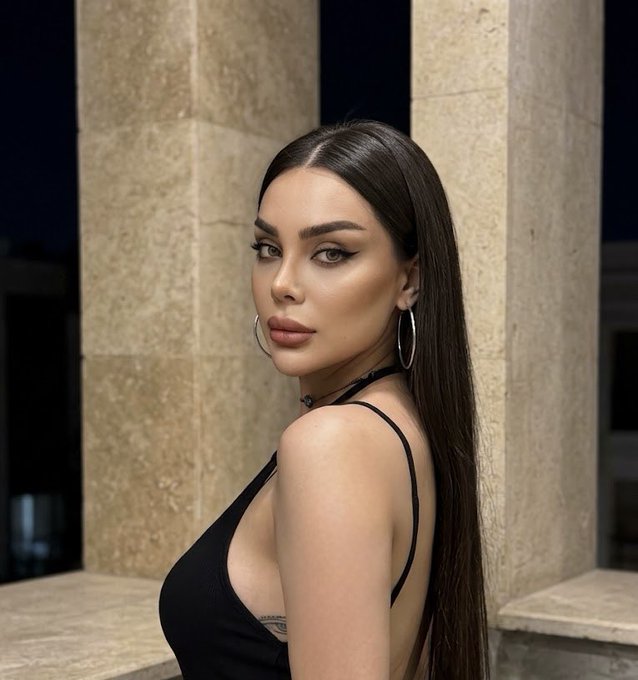 Portrait of a woman with long dark straight hair, wearing heavy makeup, large silver hoop earrings, a black tank top, and a black choker, looking over her shoulder in front of stone pillars.