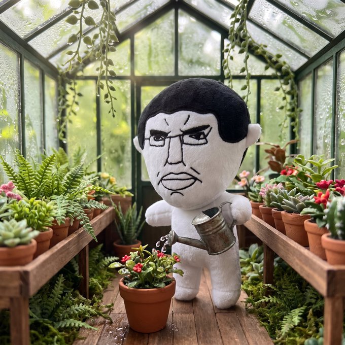 A plush doll with an angry facial expression and black hair, holding a metal watering can over a small potted flower in a greenhouse filled with various plants.