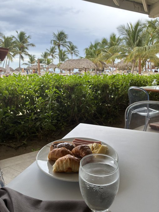 A plate of assorted pastries including croissants and slices of cake sits on a white table next to a glass of water, with a tropical resort scene featuring palm trees and thatched umbrellas in the background.