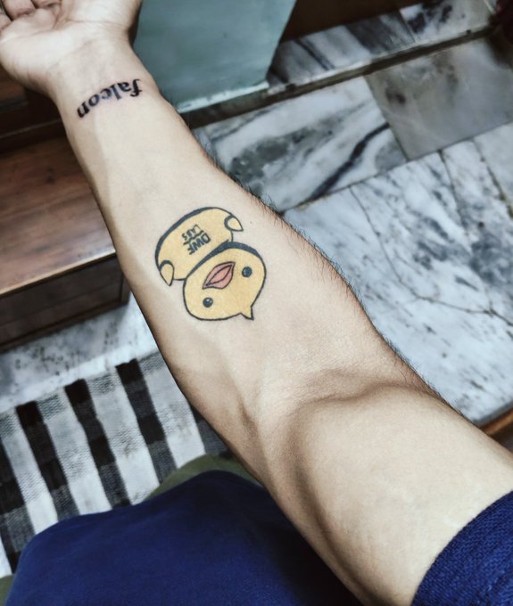 Close-up of a person's forearm displaying a small cartoon tattoo of a yellow chick/duck character, with the text "fal.com" written in cursive on the inner wrist above it.