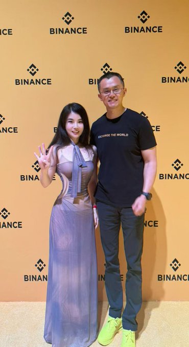 A woman in a lavender gown poses with a man wearing a "CHANGE THE WORLD" t-shirt in front of a Binance-branded backdrop, fitting the "Binance Brand Ambassador" caption as both appear as featured guests at a Binance-sponsored event.