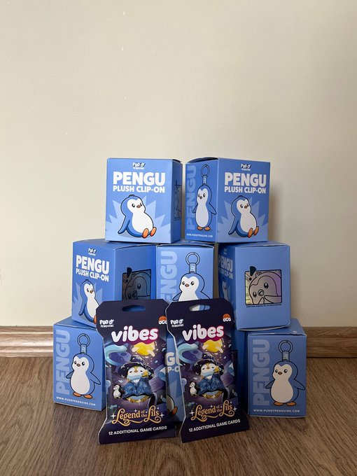 A stack of blue 'PENGU PLUSH CLIP-ON' toy boxes and two 'VIBES' Legends of the Multiverse card packs sit on a wooden floor.