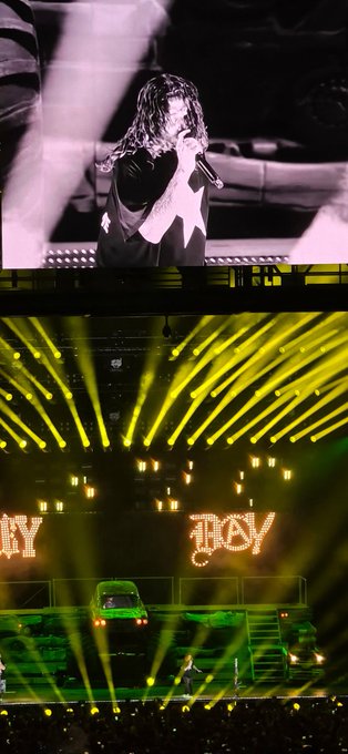 A performer sings into a microphone on stage under yellow spotlights, with a large screen showing a close-up of the artist and illuminated "HOLY" signs above a vintage car prop, capturing the lively atmosphere of the "$B" concert the user attended.