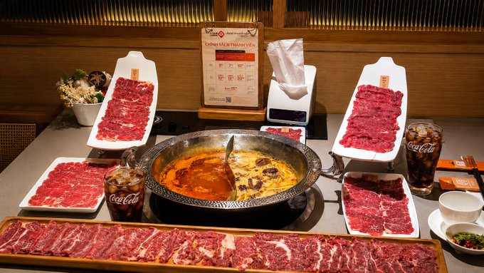 A hot pot meal featuring a divided pot with red and yellow broths, surrounded by various cuts of raw beef and a glass of Coca-Cola on a restaurant table.