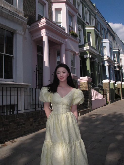 A woman in a flowing yellow dress stands on a London street lined with pastel-colored townhouses, embodying a soft, sunny aesthetic. The image pairs with the post's "GM" greeting and "Don't make Mom mad" caption, suggesting a playful maternal warmth despite the elegant, composed pose.