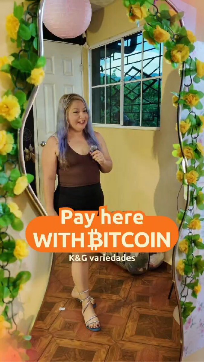 The image features a woman standing next to an archway with flowers hanging from it. The archway has a sign that says "Pay Here With Bitcoin" written on it, indicating that the shop accepts Bitcoin as payment.