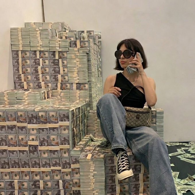A woman in a black halter top, baggy jeans, Converse sneakers, and oversized sunglasses sits on stacks of banded cash bundles while talking on a phone, with a Gucci bag at her side.