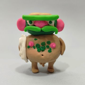 A handmade clay figurine depicts a character with a hamburger-shaped head featuring lettuce, pink ears, and a mustache, standing on a brown rounded body with green writing on its belly.