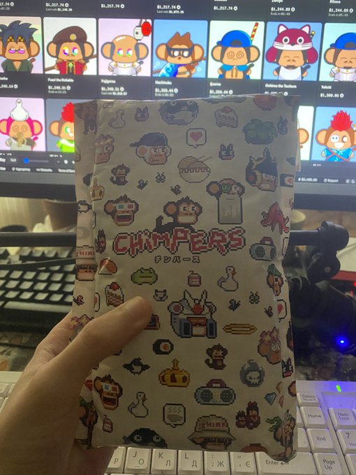 A hand holding a sealed package featuring 'CHIMPERS' pixel art characters, with a computer screen in the background displaying a collection of similar NFT monkey characters and their prices.