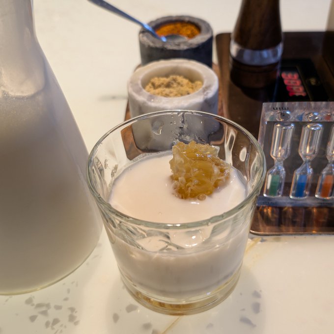 A glass contains a milky white beverage topped with a piece of honeycomb, with various bottles, containers, and colorful vials visible on the counter behind it.
