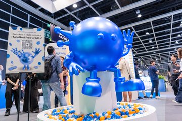 A large blue robot mascot statue with googly eyes greets visitors at the LEAP East conference, surrounded by a display of orange and blue balls and a sign reading "Take home a Mind of the Month! 1 Month of Free Coffee" alongside a QR code, capturing the playful promotional atmosphere the post describes for @hellominds_ and @Moca_Network's Air³ collaboration.