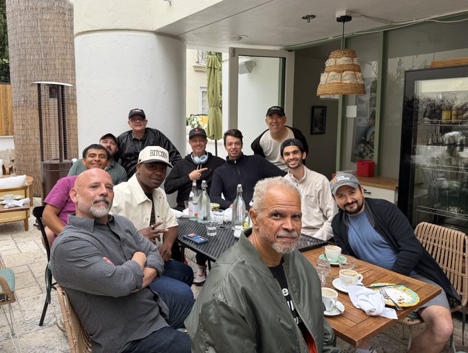 A group photo of about ten men gathered around an outdoor patio table, with the man in front wearing a green bomber jacket and another wearing a white 'BEYOND' cap.