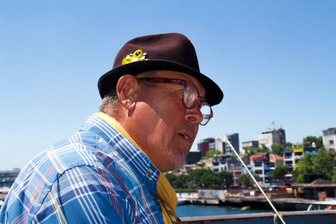 A side profile of an older man wearing a black hat with a yellow pin, glasses, and a blue plaid shirt, photographed outdoors with a cityscape in the background.