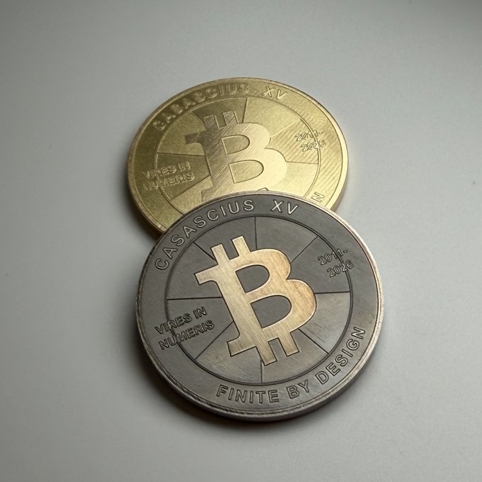 Two physical Bitcoin commemorative coins rest on a gray surface; both are engraved with 'CASASCIUS XV', 'VIRES IN NUMERIS', 'FINITE BY DESIGN', and the dates '2011-2013' and '2011-2020'.