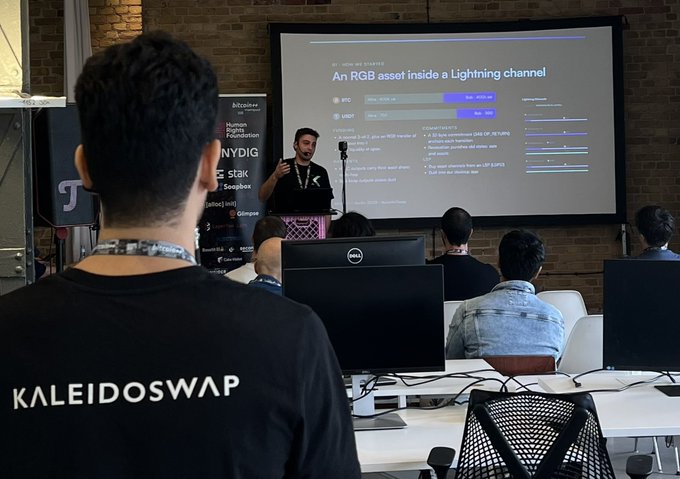 A conference setting with a speaker presenting "An RGB asset inside a Lightning channel" on a screen, while attendees sit at computers with a person in the foreground wearing a "KALEIDOSWAP" shirt.