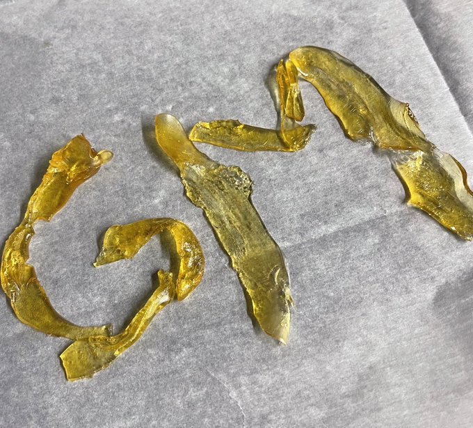 A translucent, golden-amber shatter (cannabis rosin) spread across a white paper, its brittle, glassy texture catching the light. The image aligns with the post's "Gm degens Big week ahead" caption, showcasing a celebratory morning vibe typical of cannabis community culture ahead of an anticipated week.