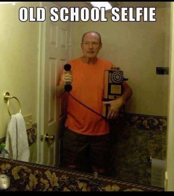 A meme image captioned 'OLD SCHOOL SELFIE' showing a man in an orange shirt taking a mirror photo while holding a vintage wall-mounted telephone receiver.