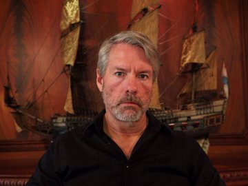 A middle-aged man with gray hair and a beard, wearing a black shirt, sits in front of an ornate wooden backdrop featuring a detailed model sailing ship. The image likely accompanies commentary or analysis related to Michael Saylor's statements comparing STRC to "kerosene" for building other financial products.