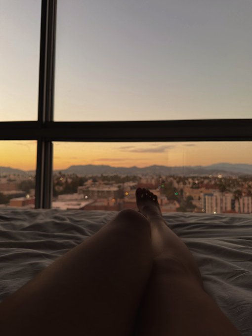 A silhouetted figure relaxes on a bed with their feet up toward a large window, overlooking a cityscape bathed in golden sunset hues with mountains in the distance. The serene, contemplative scene captures the dreamy "should I move here?" travel mood, perfectly pairing with the post's playful musing about relocating anywhere on a whim.