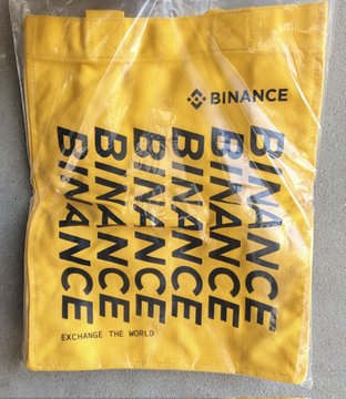 A bright yellow drawstring bag features the Binance logo repeated multiple times and the slogan 'EXCHANGE THE WORLD'.