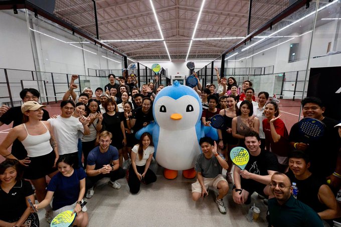 The image features a large group of people posing together for a photo, with some holding tennis rackets and others holding potted plants. The group consists of both men and women, all dressed similarly in matching outfits. They are gathered around a penguin mascot that is standing on its hind legs, surrounded by the crowd. The penguin mascot appears to be wearing a hat and has a blue body with orange feet.

The post includes text that reads: "Love seeing cross chain communities partner with each other for events. I'm a huge advocate of cross pollination and seeing two of my faves collaborate makes me so happy! Many more to come @AvaxTeam1 @pudgypenguins".