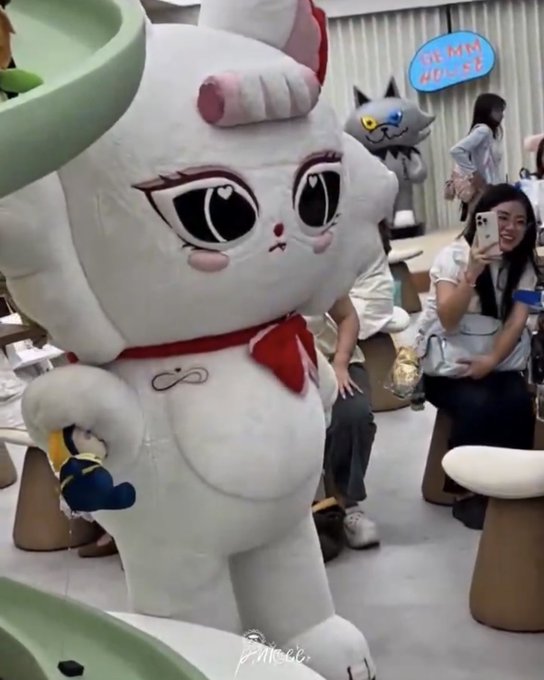 A large white cartoon character mascot with big eyes and a red bow stands in what appears to be a store, with people in the background taking photos.