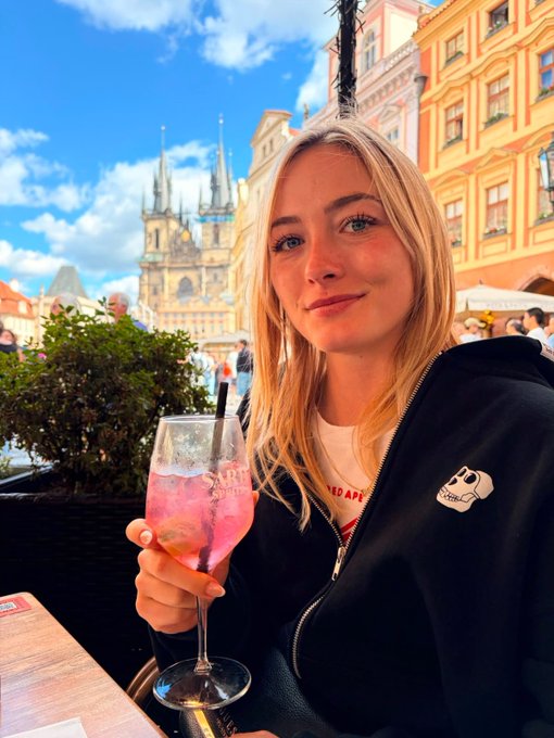 A young blonde woman smiles while holding a glass of pink Aperol Spritz at an outdoor café in Prague's Old Town Square, with the Gothic Church of Our Lady before Týn visible in the background.