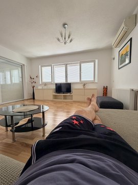 A first-person view from a couch in a modern, minimalist living room showing crossed legs in dark sweatpants featuring red star designs, with a TV, coffee table, and large windows visible in the background.