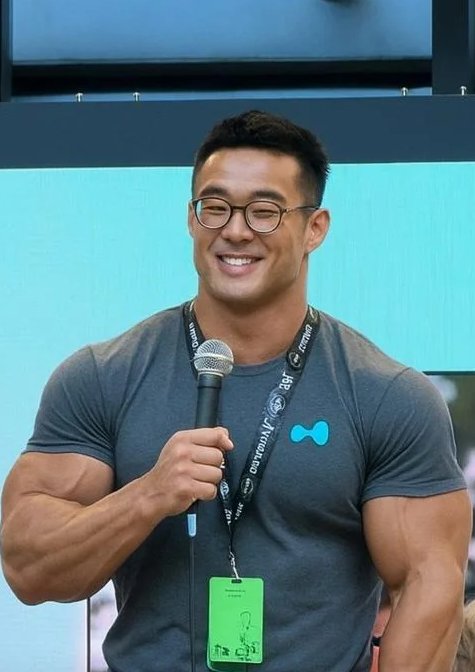 A muscular man wearing glasses and a gray T-shirt with a teal bowtie logo smiles while holding a microphone; his lanyard shows "Activision" and "BG" branding.