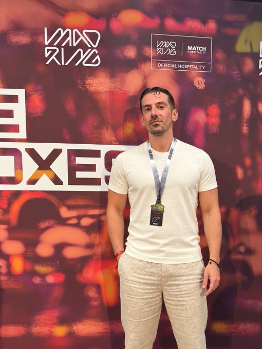 A man in a white t-shirt and linen pants wearing a lanyard, standing in front of a 'MAD RING' branded backdrop with 'OFFICIAL HOSPITALITY' and 'MATCH' signage.