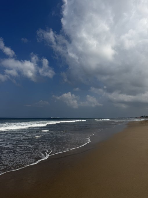 A scenic view of a sandy beach with gentle waves rolling onto the shore under a dramatic blue sky filled with large, fluffy white clouds.