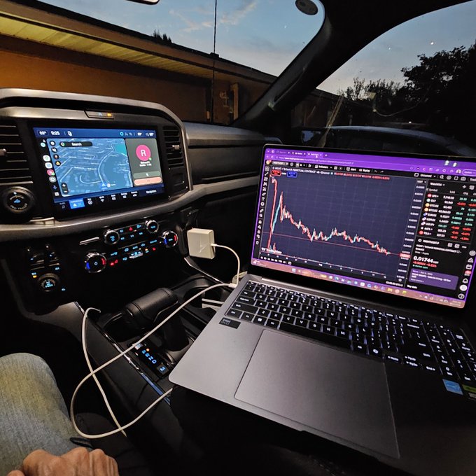 A laptop on a car's center console displaying a cryptocurrency trading chart with declining prices, connected via cable, with the car's infotainment screen showing a map navigation in the background.