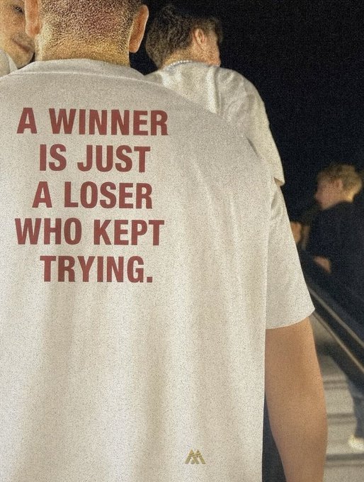 A person wears a light-colored t-shirt bearing the bold red motivational quote: 'A WINNER IS JUST A LOSER WHO KEPT TRYING.'