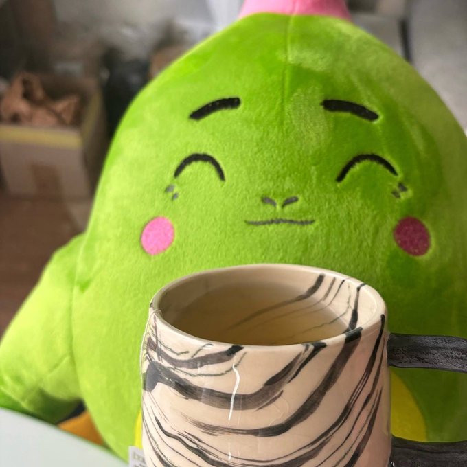 A green plush toy character with closed smiling eyes and pink cheeks sitting behind a beige and black striped ceramic mug, appearing to enjoy a drink.
