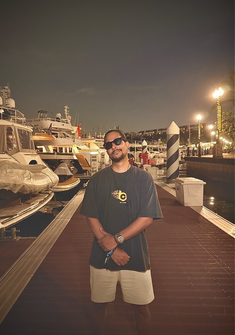 A man wearing sunglasses and a black "GN" logo t-shirt poses on a marina dock at night, with luxury yachts and illuminated harbor lights in the background, fitting the "GN Fam" theme.