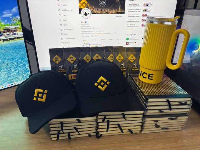 Binance merchandise, including two black caps, stacked notebooks, and a yellow tumbler, sits on a desk in front of a monitor showing a Twitter feed.