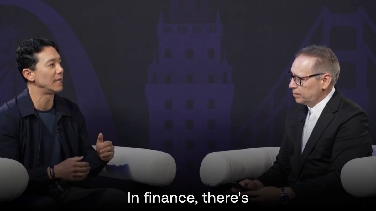 The image shows two men sitting across from each other at a table with their hands placed on the table. They appear to be engaged in conversation or discussion about finance. The background of the image features a city skyline, which suggests that they might be discussing financial matters related to urban development and planning.