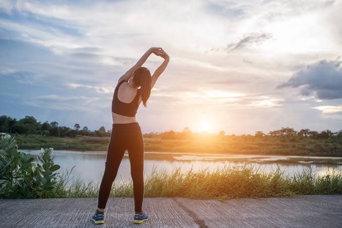 A woman in black athletic wear performing a side stretch exercise outdoors on a paved path at sunset, with a scenic view of a lake, grass, and a colorful sky in the background.