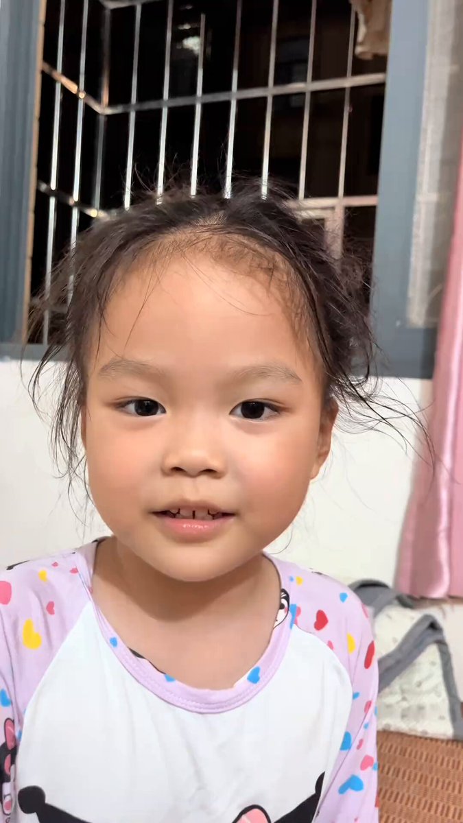 The image features a young girl with pigtails, wearing a pink shirt with white polka dots. She has dark hair and is looking directly at the camera. The background of the photo shows a window behind her, suggesting that she might be indoors or in an area with natural light coming through the window. There are also some text elements present in the image, including "现在的小孩姐:六一儿童节我要买纳斯达克 $SPY $QQQ".