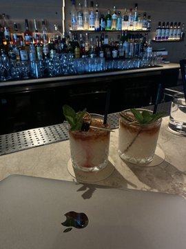Two iced cocktails garnished with mint leaves sit on a bar counter in front of a backlit shelf stocked with liquor bottles, while a Macbook laptop displaying the Apple logo rests on the counter in the foreground. The image accompanies a post about prioritizing self-worth ("Chudmaxxing") by cutting ties with someone undervalued them, suggesting a solo drink at a bar as a moment of reflection.