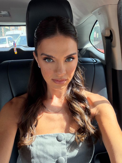 A selfie of a woman with long brown hair and blue eyes wearing a gray top, sitting inside a car.