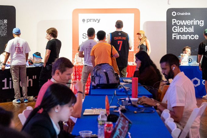 The image shows a group of people gathered around tables at an event, possibly a tech conference or meetup. They are using laptops and other devices while standing near each other. The room has a blue tablecloth covering most of the tables, with some attendees sitting on chairs placed around them. There is also a whiteboard in the background that appears to be related to the event's theme.

The post features an image of a person wearing an orange shirt and a name tag that reads "@privy_io". The caption explains how Privy helps developers build seamless onboarding experiences for users, which can include embedded wallets, account management, and user authentication. This information is relevant to the event as it suggests that Privy's technology might be being showcased or discussed at this gathering.
