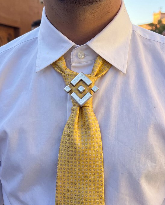 Close-up photograph of a man wearing a white dress shirt with a yellow patterned tie featuring a Binance (BNB) cryptocurrency logo tie clip/marker in gold.