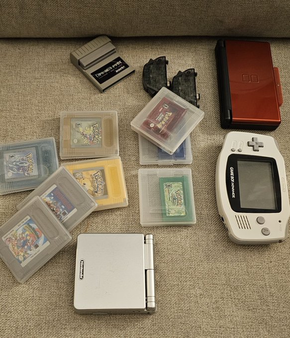 A collection of retro gaming items arranged on carpet, including several Game Boy game cartridges (Pokémon), a Game Boy Advance SP, a Nintendo DS, and what appears to be a copy of Tetris.