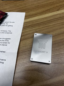 A metallic plate engraved with the number '1000' and 'ALOHACK' lies on a wooden table next to a printed document and a red and black marker.
