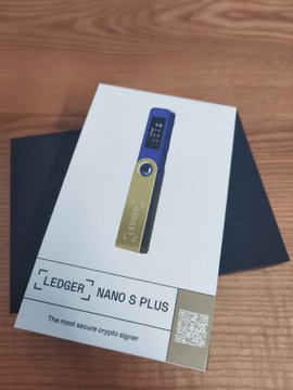 The image shows a small box with a blue and gold design, sitting on top of a wooden surface. The box appears to be a Ledger Nano S Plus wallet, which is described as "the most secure crypto signer."