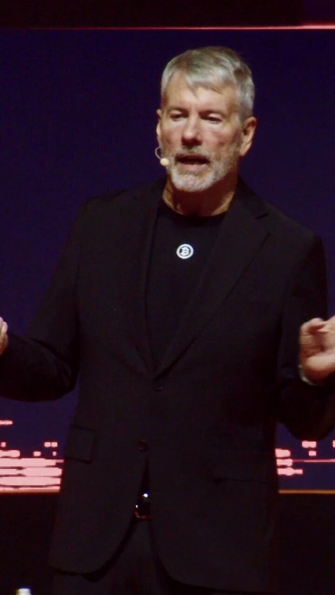 The image features an older man wearing a black suit, standing in front of a large screen with his hands outstretched as if he's giving a speech or presentation. The background includes a purple wall and a red light, which could be part of a stage setup for an event. The caption reads "This is how Michael Saylor plans to bring Bitcoin to a billion people sooner than we expect". This suggests that the man in the suit might be discussing his vision for the widespread adoption of Bitcoin or sharing insights about its potential impact on society and economy.